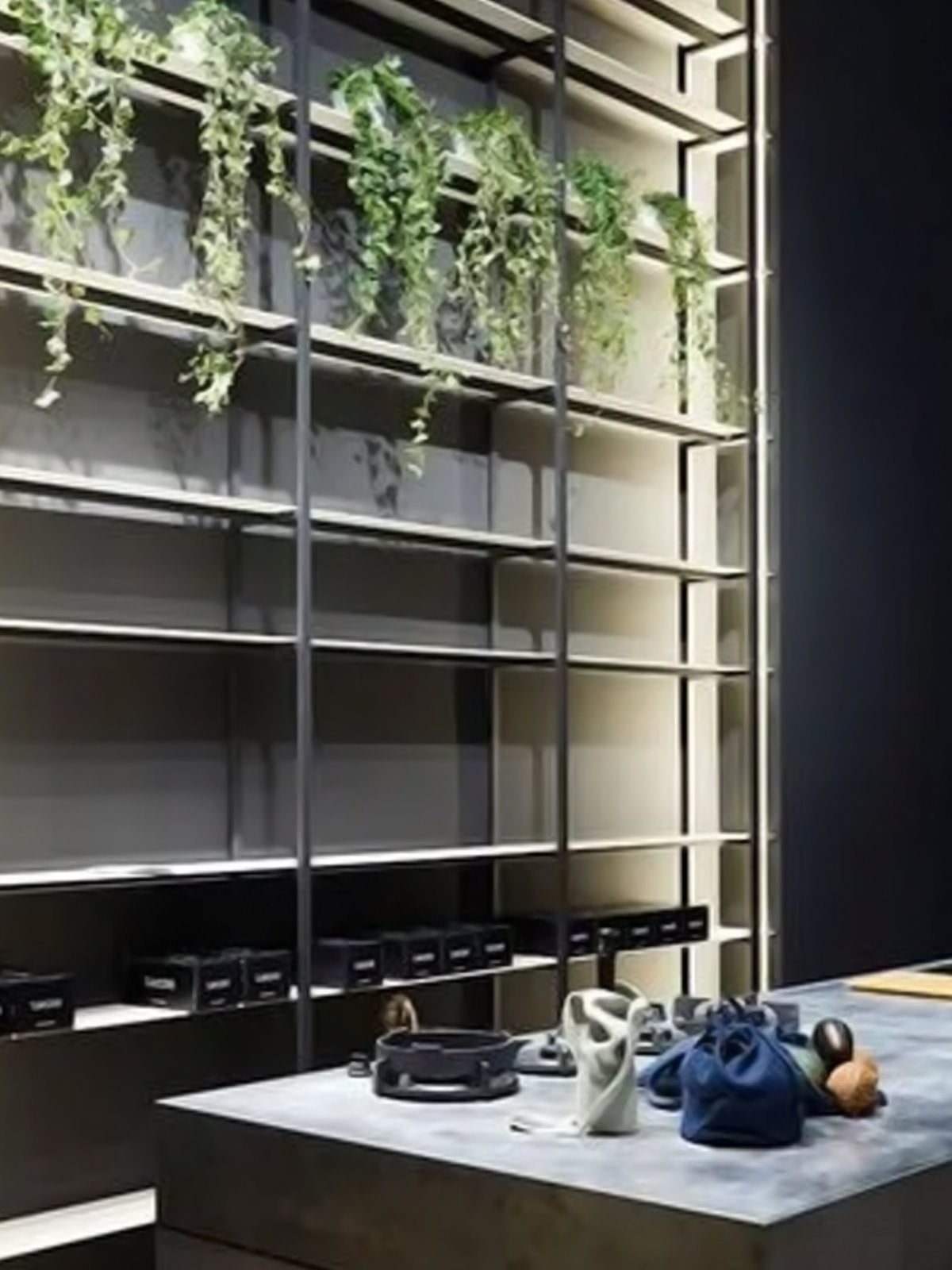 Ein modernes Verkaufsdisplay mit einem Metallregal, das oben grüne Pflanzen und unten mehrere schwarze Kisten enthält. Im Vordergrund werden auf einer Betontheke Accessoires und kleine Taschen ausgestellt.
