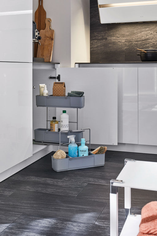 Geöffneter Unterschrank mit herausgezogenem Putzmittelauszug. Auf dem Fußboden daneben ein herausnehmbarer Korb mit Küchen- und Reinigungsutensilien.