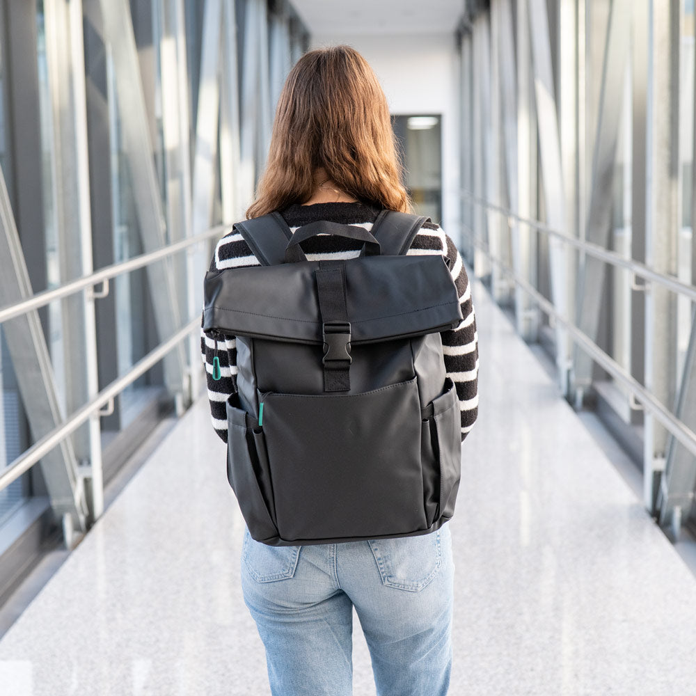 Eine Person mit langen braunen Haaren, einem schwarz-weiß gestreiften Pullover und hellblauen Jeans steht auf einem gläsernen Steg mit einem Kesseböhmer-Rucksack - einem geräumigen schwarzen Rucksack, der sich ideal zum Transport von Mahlzeiten oder einer Brotdose eignet. Das Foto ist von hinten aufgenommen.