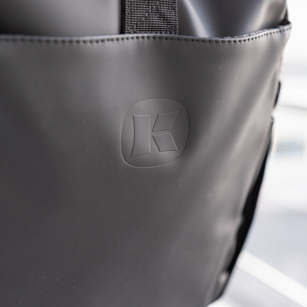 Nahaufnahme des Kesseböhmer Rucksacks in mattem Schwarz mit dezent geprägtem K-Logo auf der Vordertasche. Die sichtbare Struktur und die Nähte unterstreichen seine Qualität - ideal für den Transport von Mahlzeiten zum Mitnehmen.