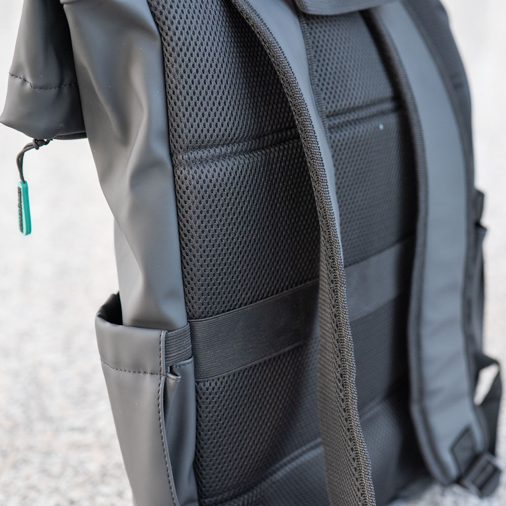 Nahaufnahme des Kesseböhmer Rucksacks in schwarz mit gepolstertem Netzrücken und breiten Trägern, ideal für Mahlzeiten to go. In die Seitentasche passt eine Kesseböhmer Brotdose, und ein Reißverschluss mit grünem Etikett sticht auf dem unscharfen grauen Grund hervor.