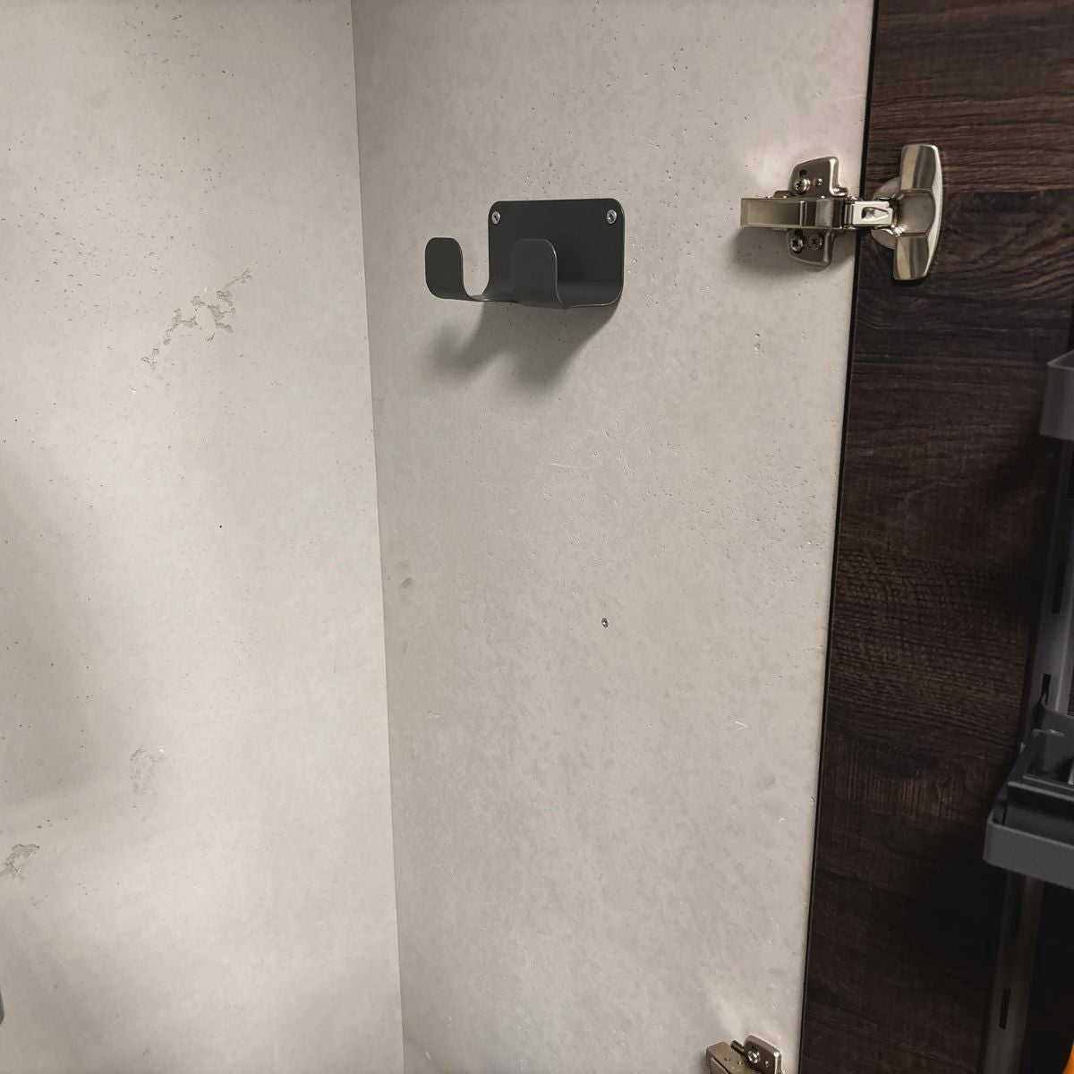 Der K-Line Seitenschlauchhalter, ein Doppelhaken aus Metall, ist an der Innenwand eines offenen Holzschranks angebracht, wobei die Tür geöffnet ist, um das Scharnier und die Innenfläche zu zeigen.