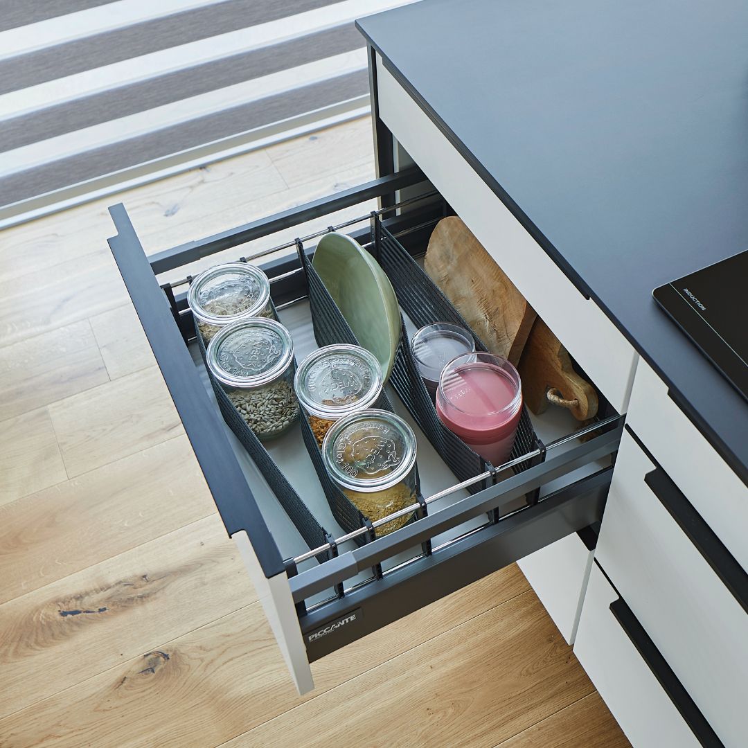 Eine offene Küchenschublade, die mit dem SpaceFlexx® Schubladen Organizer organisiert wurde, enthält Gläser mit Lebensmitteln, einen grünen Teller, ein hölzernes Schneidebrett, ein Glas mit rosa Flüssigkeit und eine durchsichtige Schüssel. Zur modernen Einrichtung gehören Nesttex Hightech-Textil-Gurtbänder und ein heller Holzfußboden.