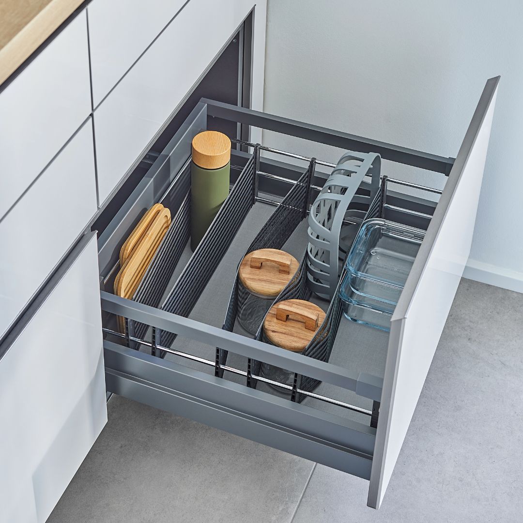 Eine offene Küchenschublade, die mit dem SpaceFlexx® Schubladen Organizer organisiert wurde, zeigt Schneidebretter, Glasbehälter mit Holzdeckeln, eine grüne Flasche und ein Geschirrregal mit nesttex Hightech-Textil-Gurtbändern in einer modernen, minimalistischen Küche.