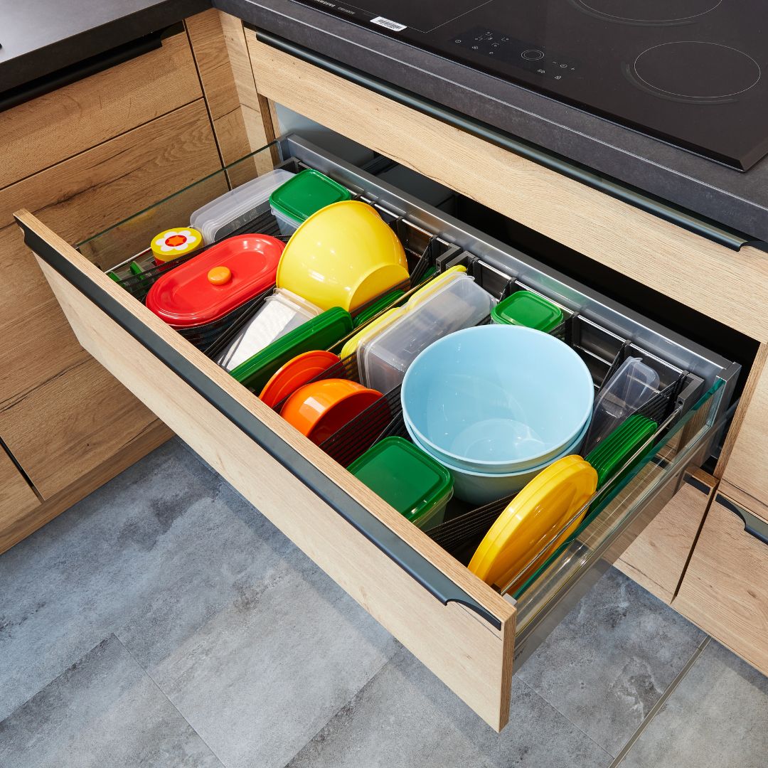 Eine Küchenschublade mit farbenfrohen Behältern, Schalen und Deckeln - darunter einige mit Hightech-Textil-Gurtbändern von nesttex aus Deutschland -, die im SpaceFlexx® Schubladen Organizer unter einer schwarzen Herdplatte und Holzschränken angeordnet sind.