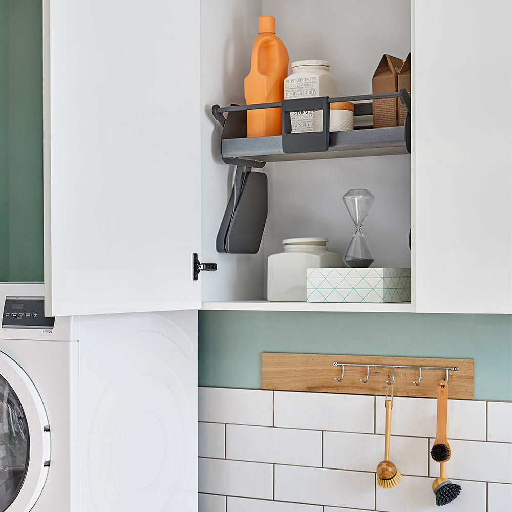 In dem offenen weißen Schrank befindet sich das iMove 60er single Auszugsystem für den Hängeschrank mit organisierten Gegenständen, Flaschen, Kisten und einer Sanduhr. Unten ist eine Waschmaschine teilweise sichtbar, über den gefliesten Wänden hängen Reinigungsbürsten für den schnellen Zugriff.