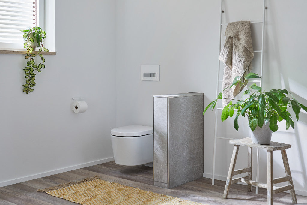 Minimalistisches Badezimmer mit einer wandmontierten Toilette, einem Handtuchhalter mit beigen Handtüchern, Topfpflanzen, einem Holzhocker, einem gelben Teppich und Licht, das durch ein Fenster einfällt.