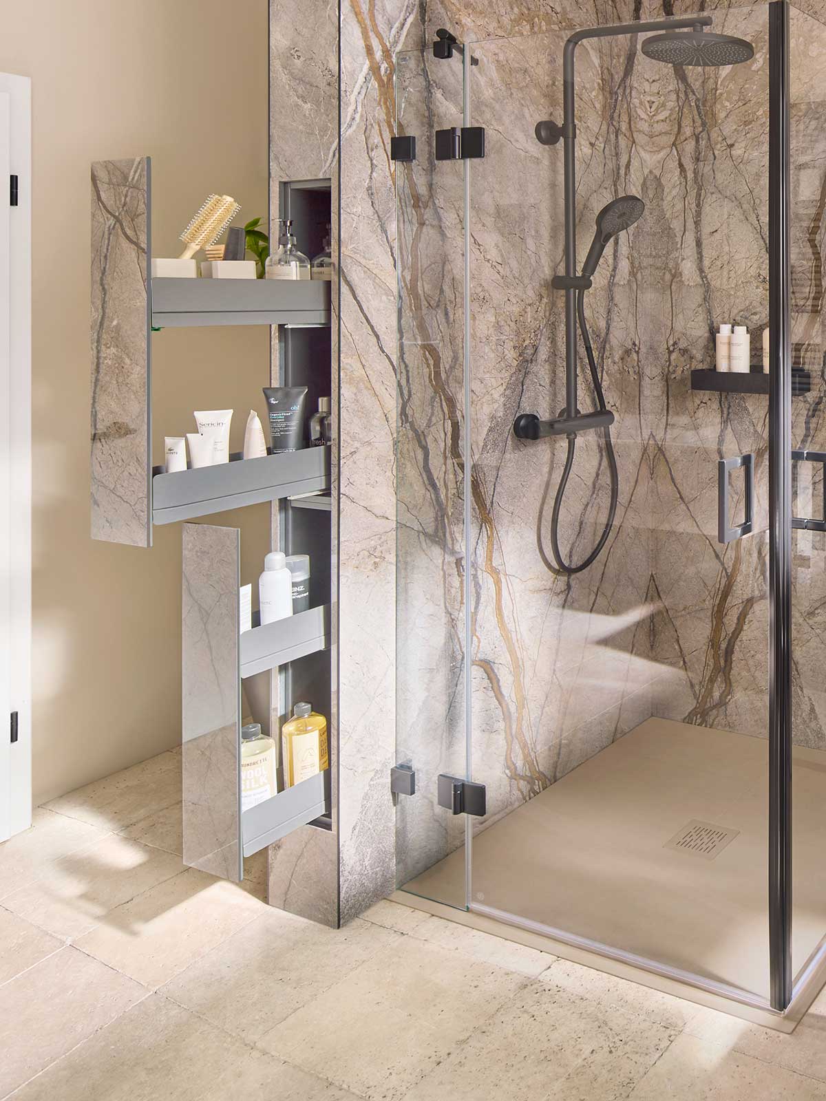 Ein modernes Badezimmer verfügt über eine verglaste Dusche mit einem Regenduschkopf. Daneben befindet sich ein hoher Spiegelschrank mit ausziehbaren Regalen, in dem verschiedene Toilettenartikel und Hautpflegeprodukte aufbewahrt werden. Die Wände sind mit Fliesen in Marmoroptik versehen.