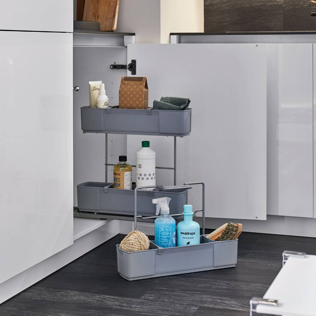 Ein cleaningAGENT - Putzschrank Organizer & Putzmittelauszug mit drei grauen Einlegeböden ist in einem weißen Küchenschrank installiert, in dem Putzmittel, Flaschen, eine Bürste und Bindfaden ordentlich aufbewahrt werden. Die offene Tür zeigt den praktischen Organizer.