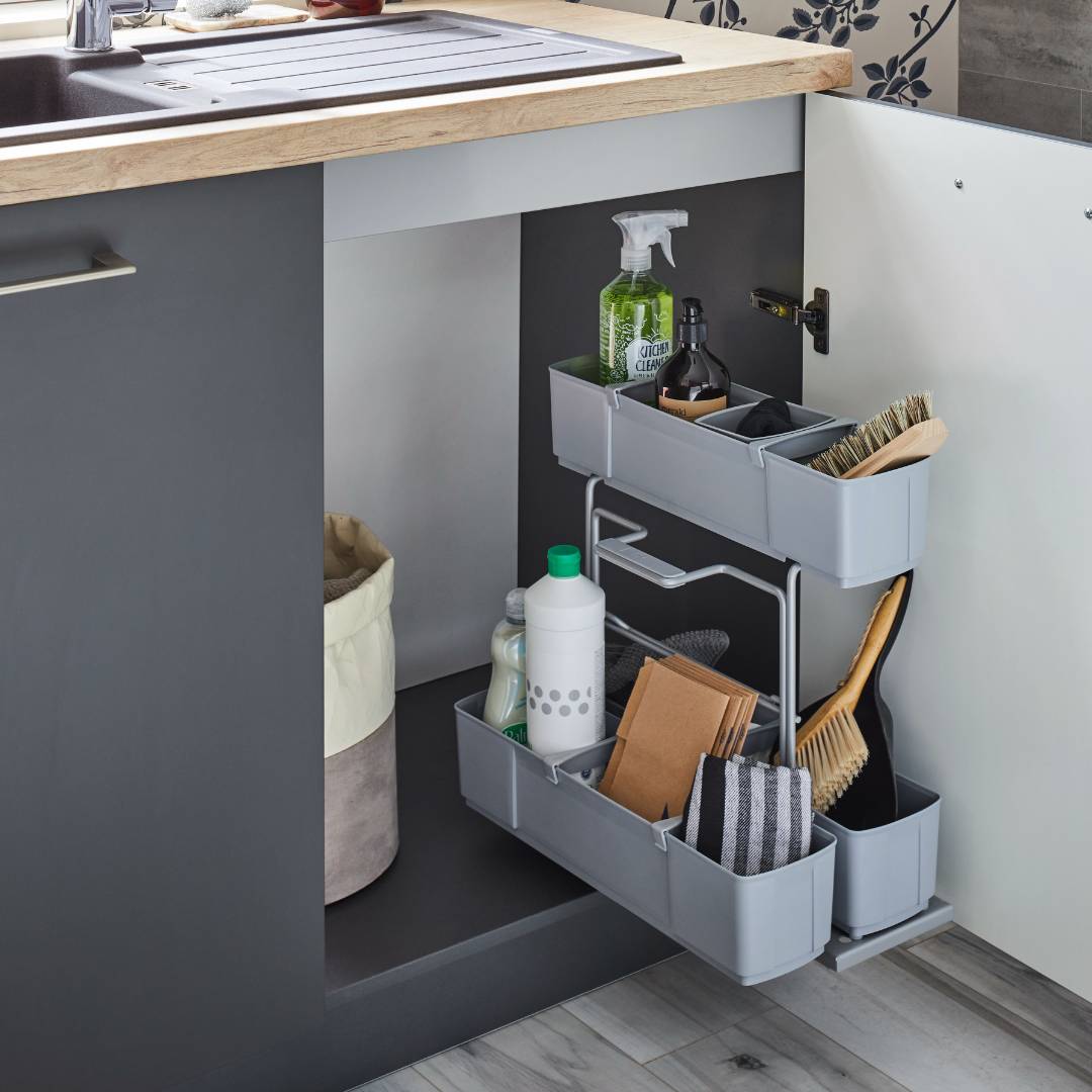 Der cleaningAGENT - Putzschrank Organizer & Putzmittelauszug mit Putzmitteln, Bürsten, Spülmittel und Tüchern steht in einem offenen Unterschrank. Auf der linken Seite des Schrankbodens befindet sich ein beiger Mülleimer.