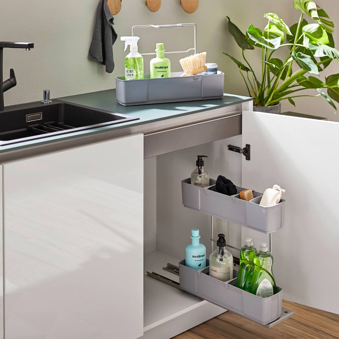 Eine moderne Küchenspüle ist mit dem cleaningAGENT - Putzschrank Organizer & Putzmittelauszug ausgestattet, der Sprühflaschen, Seife und Pinsel übersichtlich auf der Arbeitsplatte und in einem ausziehbaren Schrankregal verstaut. In der Nähe steht eine Grünpflanze.