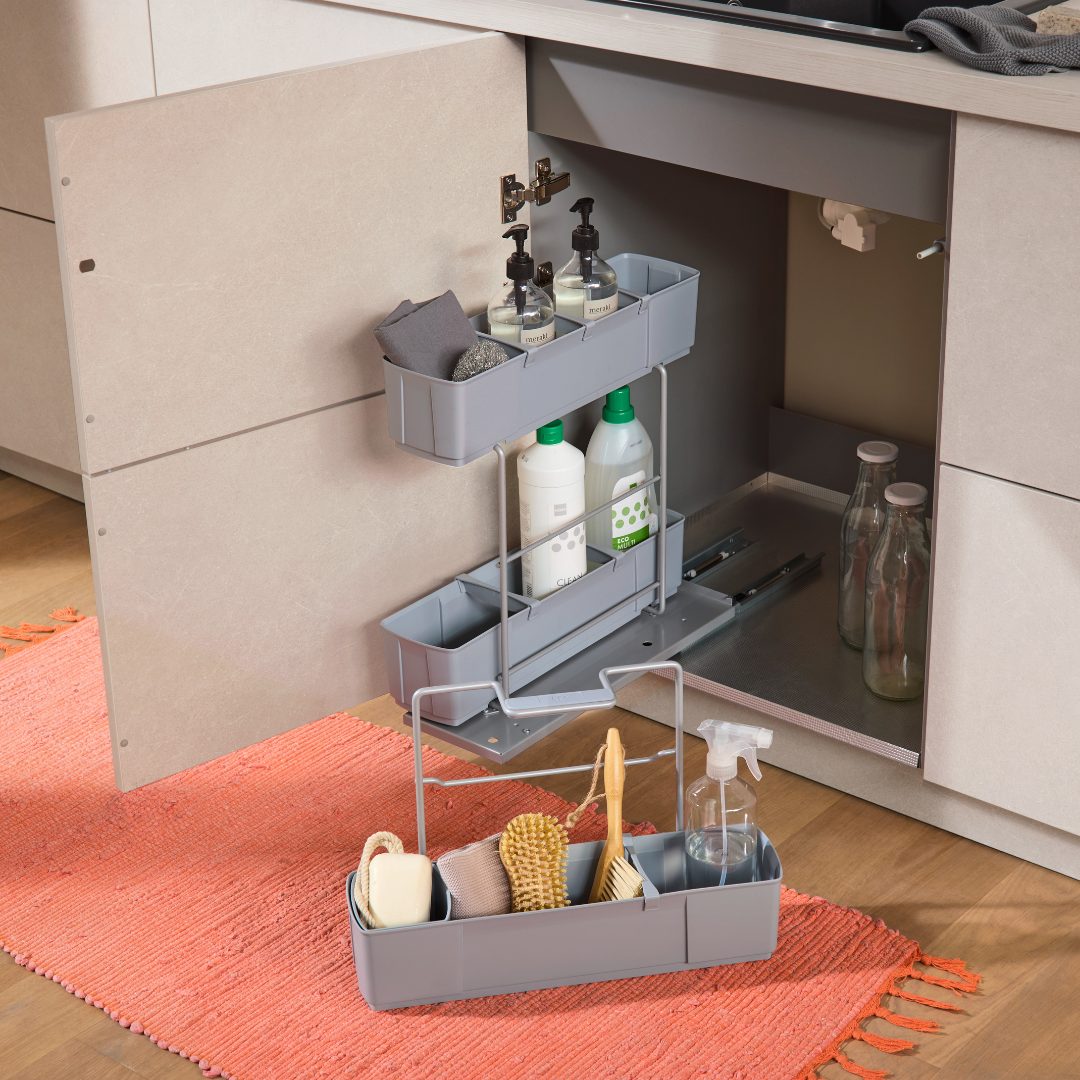 Unter dem Waschbecken befindet sich ein grauer cleaningAGENT Putzschrank Organizer & Putzmittelauszug, in dem Putzmittel und Sprühflaschen ordentlich aufbewahrt werden. Die Schranktür ist offen und gibt den Blick auf weitere Bürsten und Reinigungsmittel frei. Ein korallenroter Teppich hellt den Boden auf.