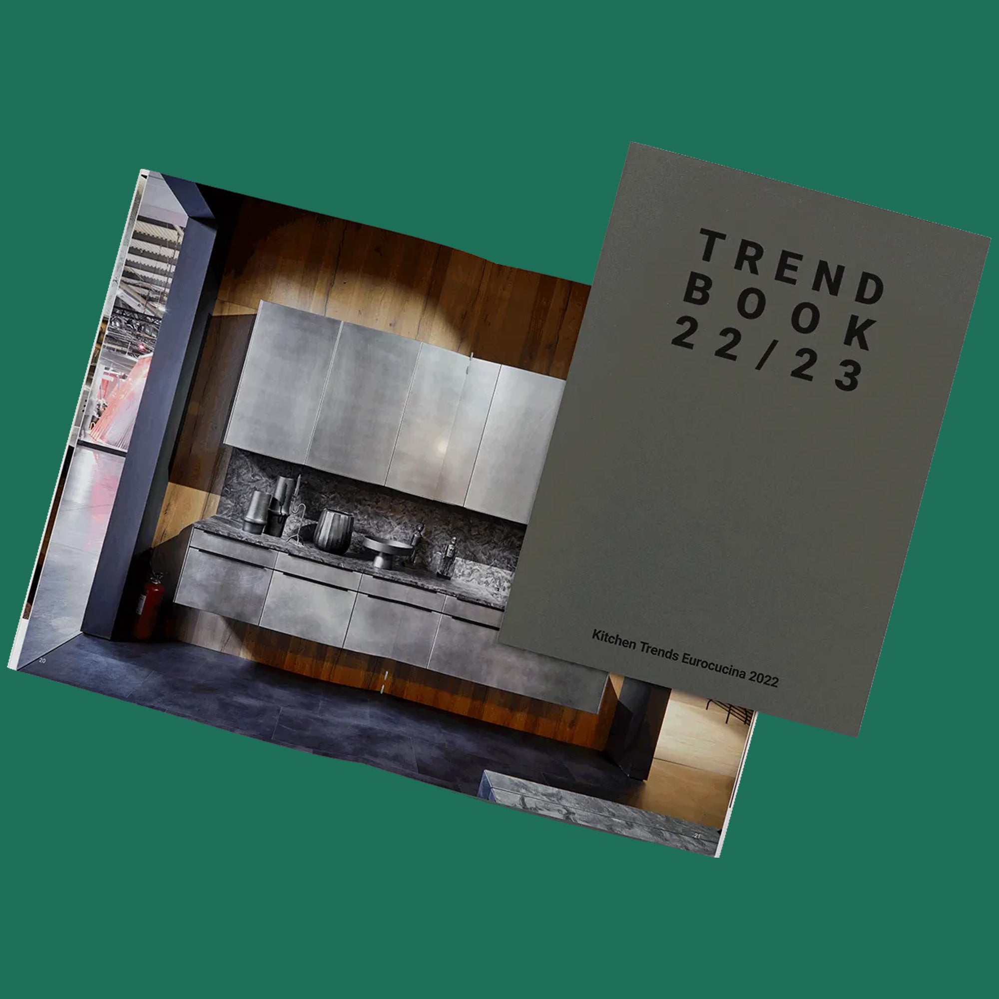 Ein graues Heft mit dem Titel TREND BOOK 22/23 liegt auf einem aufgeschlagenen Magazin, das eine moderne Küche mit glatten Metallschränken und zeitgenössischem Dekor zeigt. Der Hintergrund ist einfarbig grün.