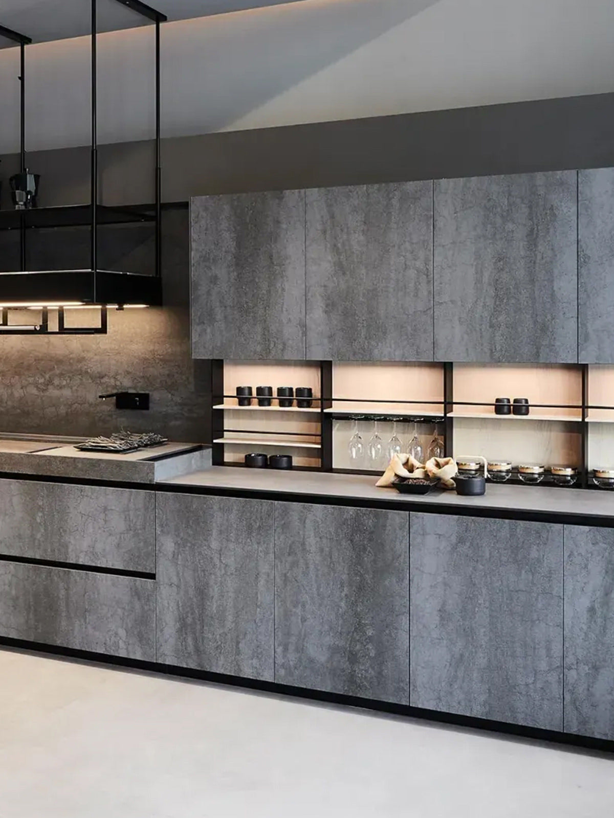 Moderne Küche mit glatten, grauen Steinschränken, offenen Regalen mit Gläsern und Geschirr, einer minimalistischen Arbeitsplatte und einem eingebauten Herd unter schwarzen Hängelampen.