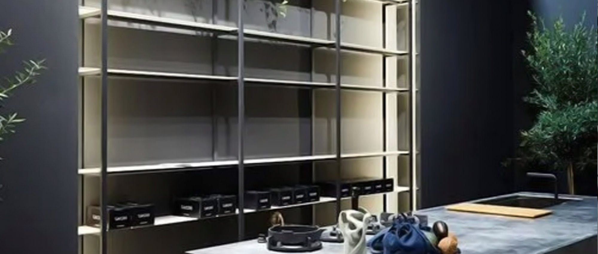 Eine moderne Küche mit einem großen, leeren Regal vor einer dunklen Wand, einer Marmorinsel im Vordergrund mit Küchenzubehör und grünen Topfpflanzen an beiden Seiten.