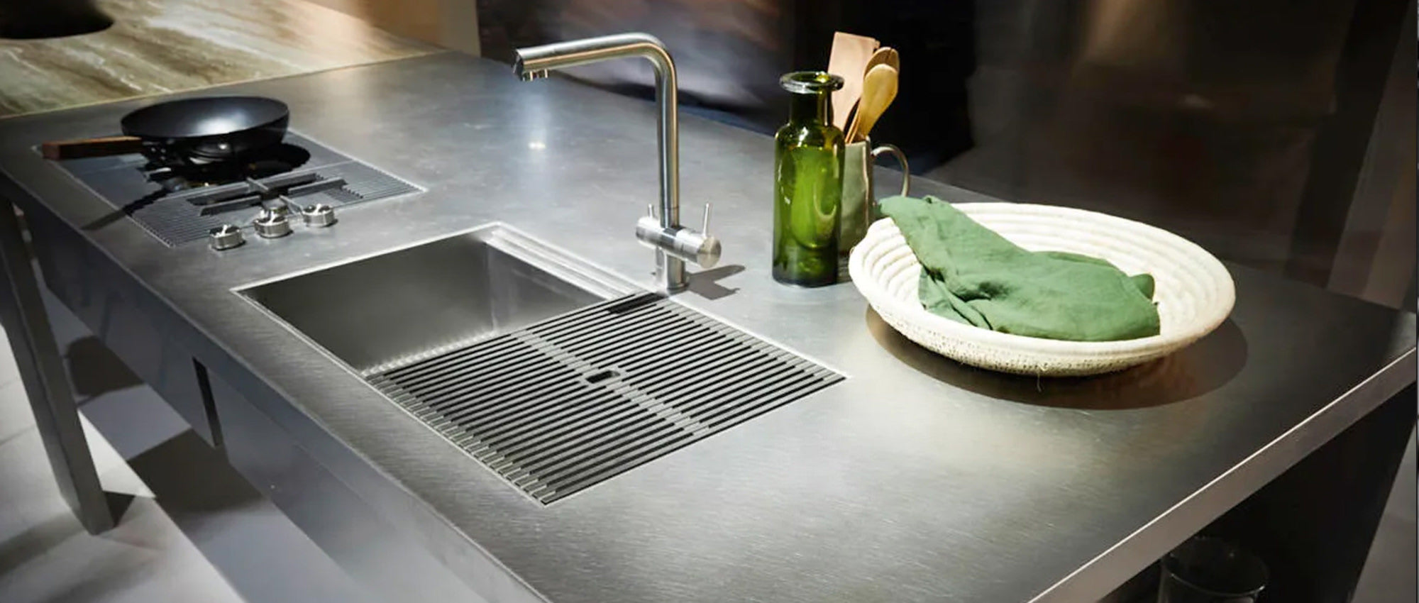 Eine moderne Küchentheke aus Edelstahl mit integrierter Spüle, Wasserhahn, Herdplatte, zwei grünen Glasflaschen, einer Bratpfanne und einem gewebten Tablett mit einem grünen Tuch.