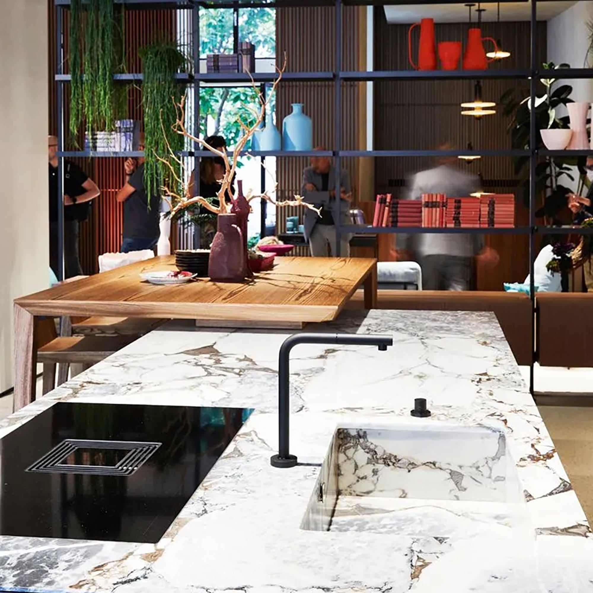Eine moderne Küche mit einer Insel aus weißem Marmor, schwarzem Wasserhahn und Spülbecken. Im Hintergrund ein hölzerner Esstisch mit dekorativen Ästen und ein stilvolles Regal mit Vasen und Büchern, in dessen Nähe sich Menschen treffen.