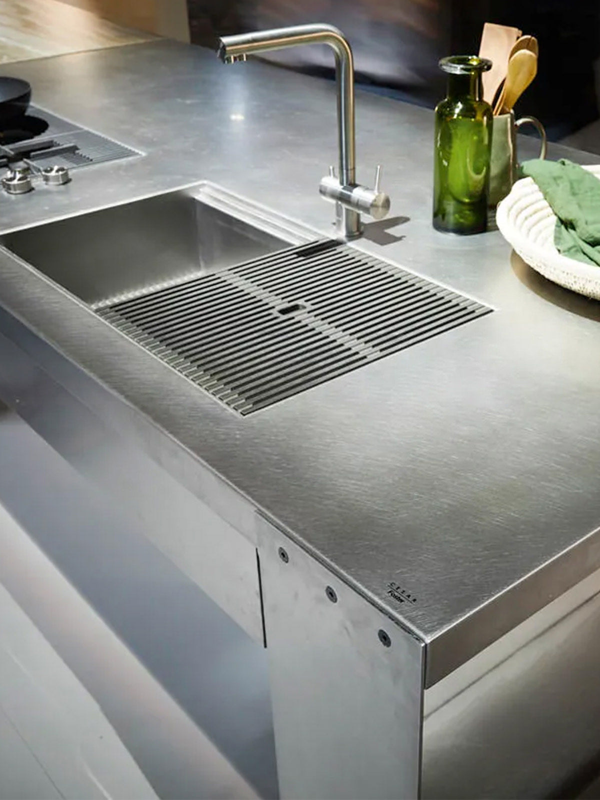 Eine moderne Edelstahlspüle mit Gitterabdeckung, ein eleganter Wasserhahn und verschiedene Küchenutensilien wie grüne Glasflaschen, Utensilien und eine Schüssel auf der Arbeitsplatte.