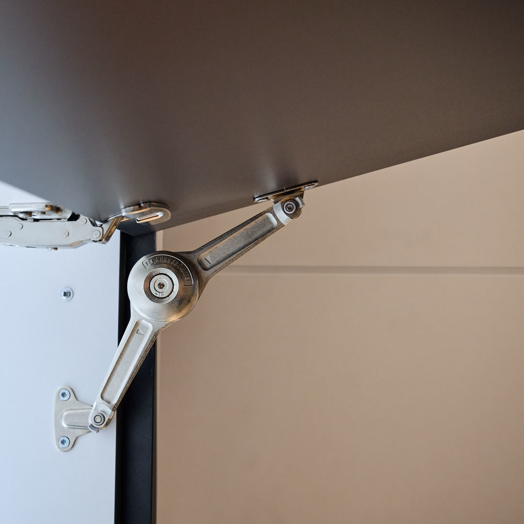 Nahaufnahme eines Scharniermechanismus für Schranktüren mit einem Metallarm und Gelenken, die an der Innenseite eines Schranks und der Tür befestigt sind und ein reibungsloses Öffnen und Schließen ermöglichen. Neutraler Hintergrund.