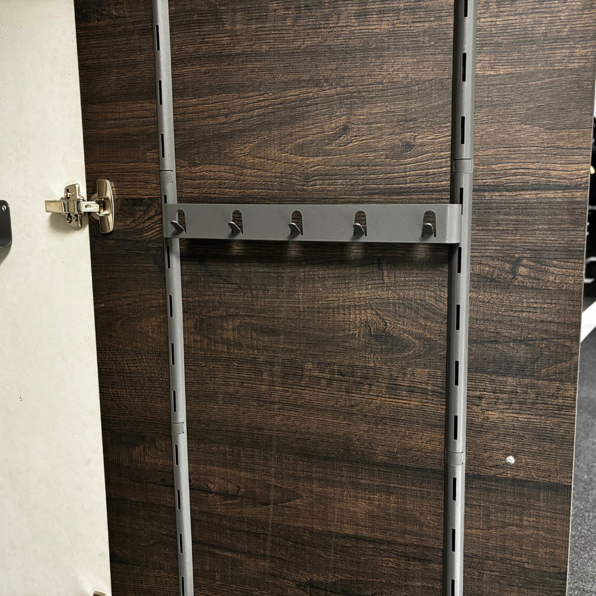 Eine Schranktür aus Holz mit dunkler Maserung öffnet sich und zeigt eine seitliche Hakenleiste von K-Line - 5 Haken, vertikal montiert als Teil eines modularen Ordnungssystems. Das Scharnier ist auf der linken Seite sichtbar.