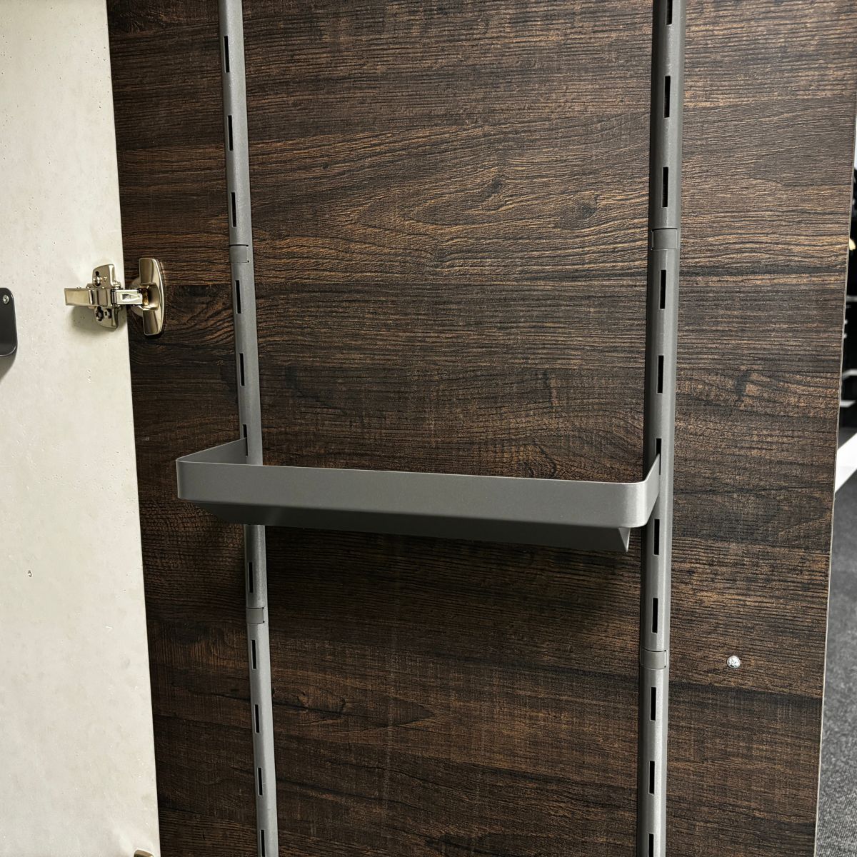 Die K-Line seitliche Papierrollenablage ist eine Metallschiene, die horizontal zwischen zwei vertikalen Schienen auf einer dunklen Holztür montiert ist. Sie hat ein verstellbares Scharnier auf der linken Seite - ideal für die Organisation der Küche.