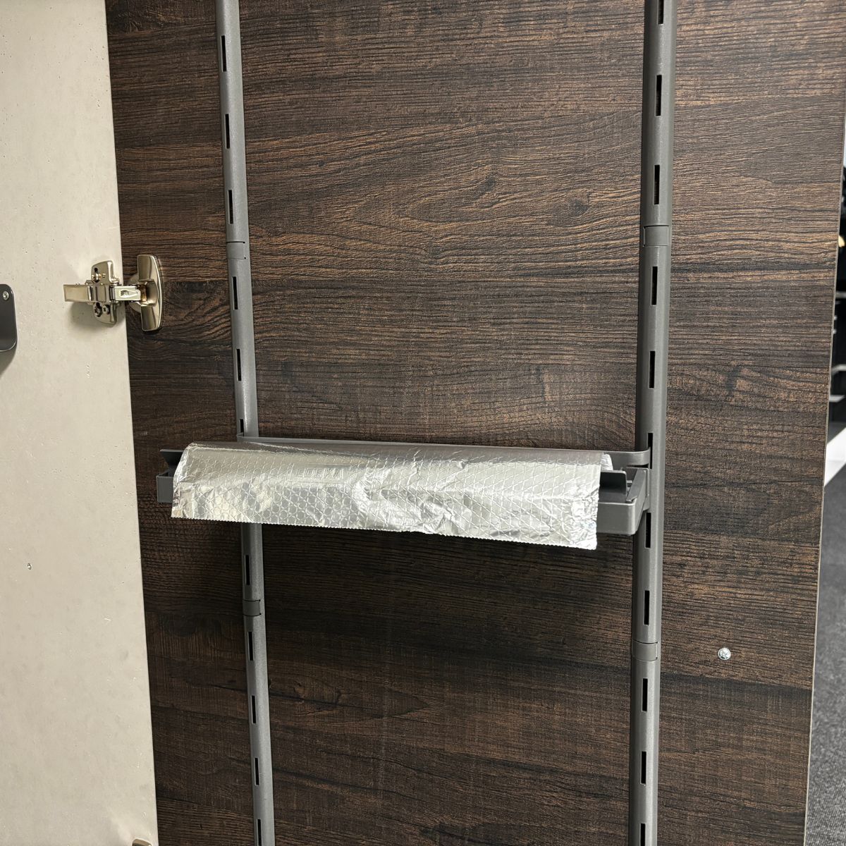 Die K-Line Seitenablage ROLLINERO - Rollenablage, ideal für die Aufbewahrung von Folie oder Frischhaltefolie, ist waagerecht zwischen zwei grauen Bügeln auf einer dunkel getäfelten Fläche mit Regalschlitzen montiert. An der linken Kante befindet sich ein Schrankscharnier.