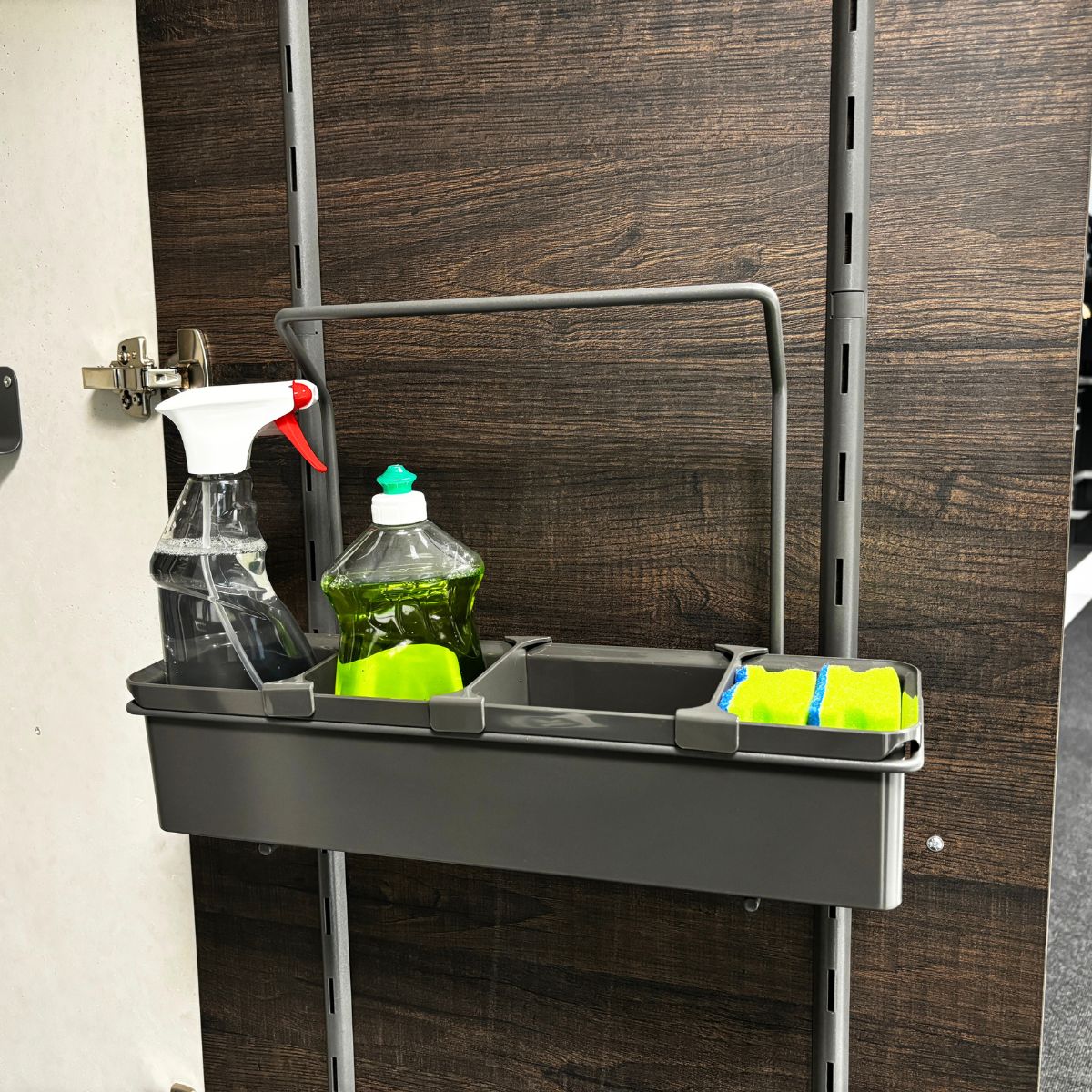 Eine an der Wand montierte graue K-Line Side Tragebox enthält eine Sprühflasche, eine Spülmittelflasche, einen grünen Schwamm und einen blau-gelben Scheuerschwamm zur geordneten Aufbewahrung von Reinigungsmitteln vor einem Hintergrund aus dunklen Holzpaneelen.