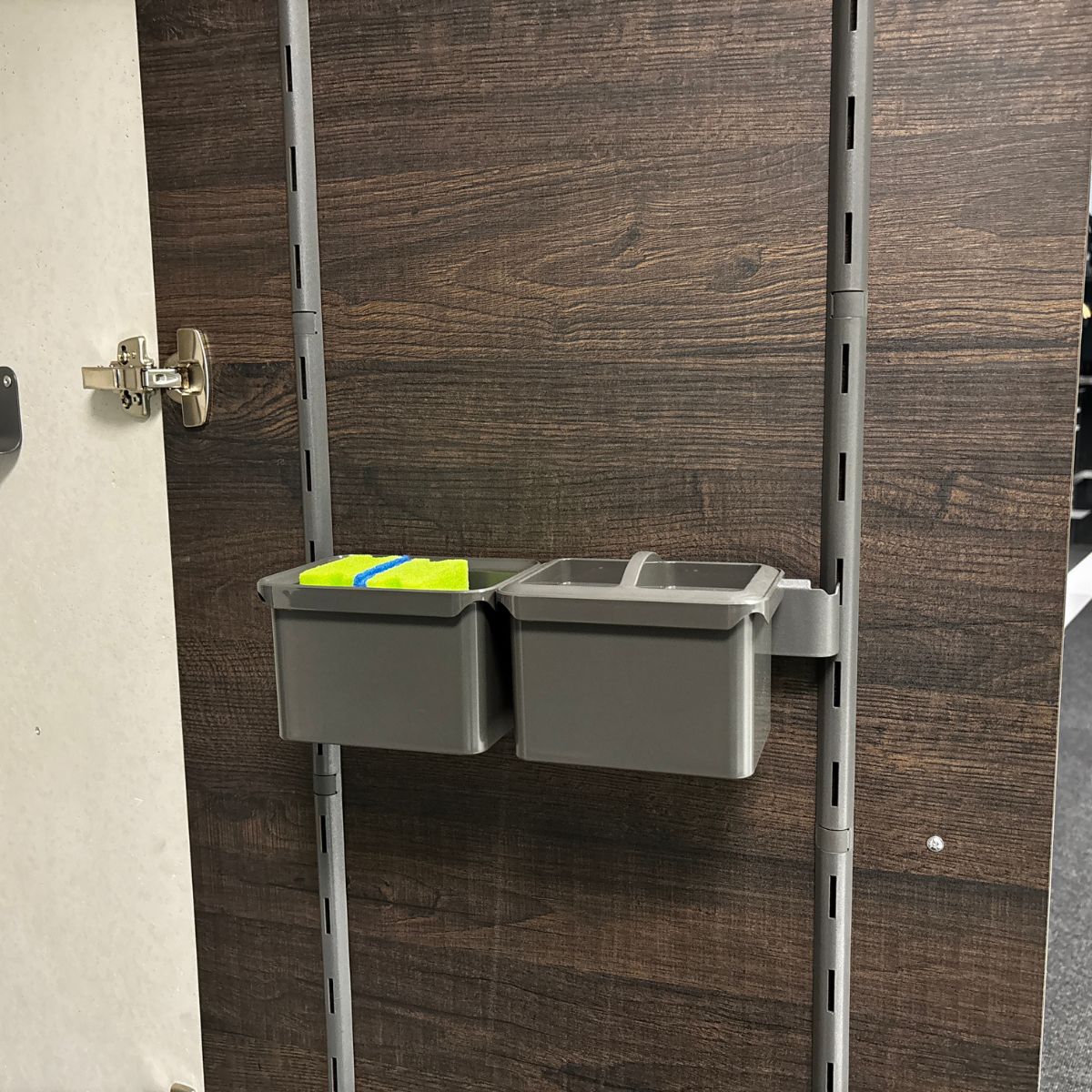 Zwei graue K-Line Seitenträgerleisten mit zwei Boxen sind auf einer Schiene an einer dunklen Holzplatte montiert. Eine Box fasst zwei gelbe und grüne Schwämme, die andere ist leer und dient der praktischen Aufbewahrung. Auf der linken Seite ist ein Schrankscharnier zu sehen.