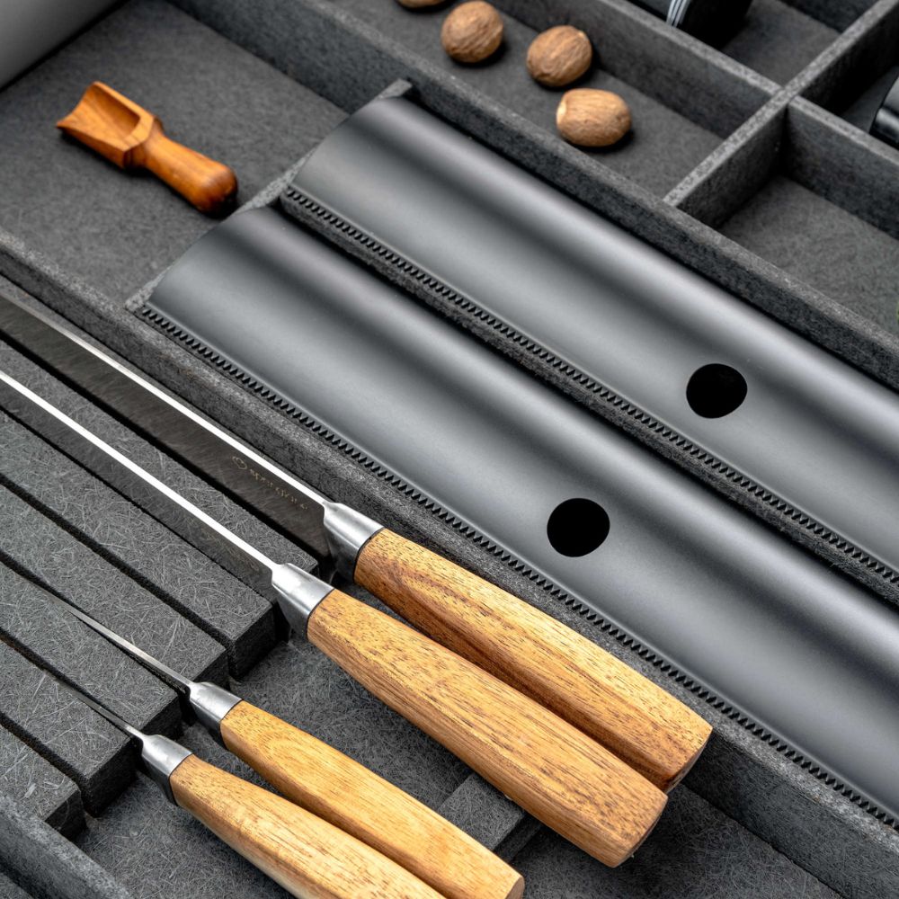 Vier Küchenmesser mit Holzgriffen sind in einem grauen Schubladenorganizer neben einer schwarzen Messerscheide, dem FineLine LiniQ Folienhalter für Frischhaltefolie, einem kleinen Holzwerkzeug und ganzen Nüssen angeordnet - perfekt für die moderne Küche.