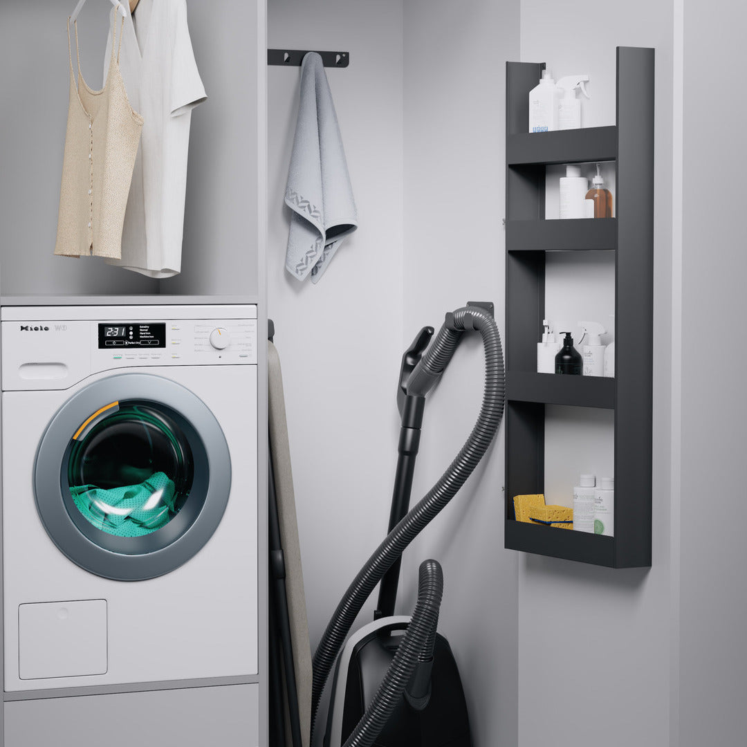 Die kompakte Waschküche ist mit einer Frontlader-Waschmaschine, einer beigen Arbeitsplatte, einem grauen Handtuchhaken, einem Staubsauger und einem an der Wand befestigten schwarzen K-Line Side EasyRack ausgestattet, das Reinigungsmittel für eine intelligente Hochschrankorganisation bereithält.