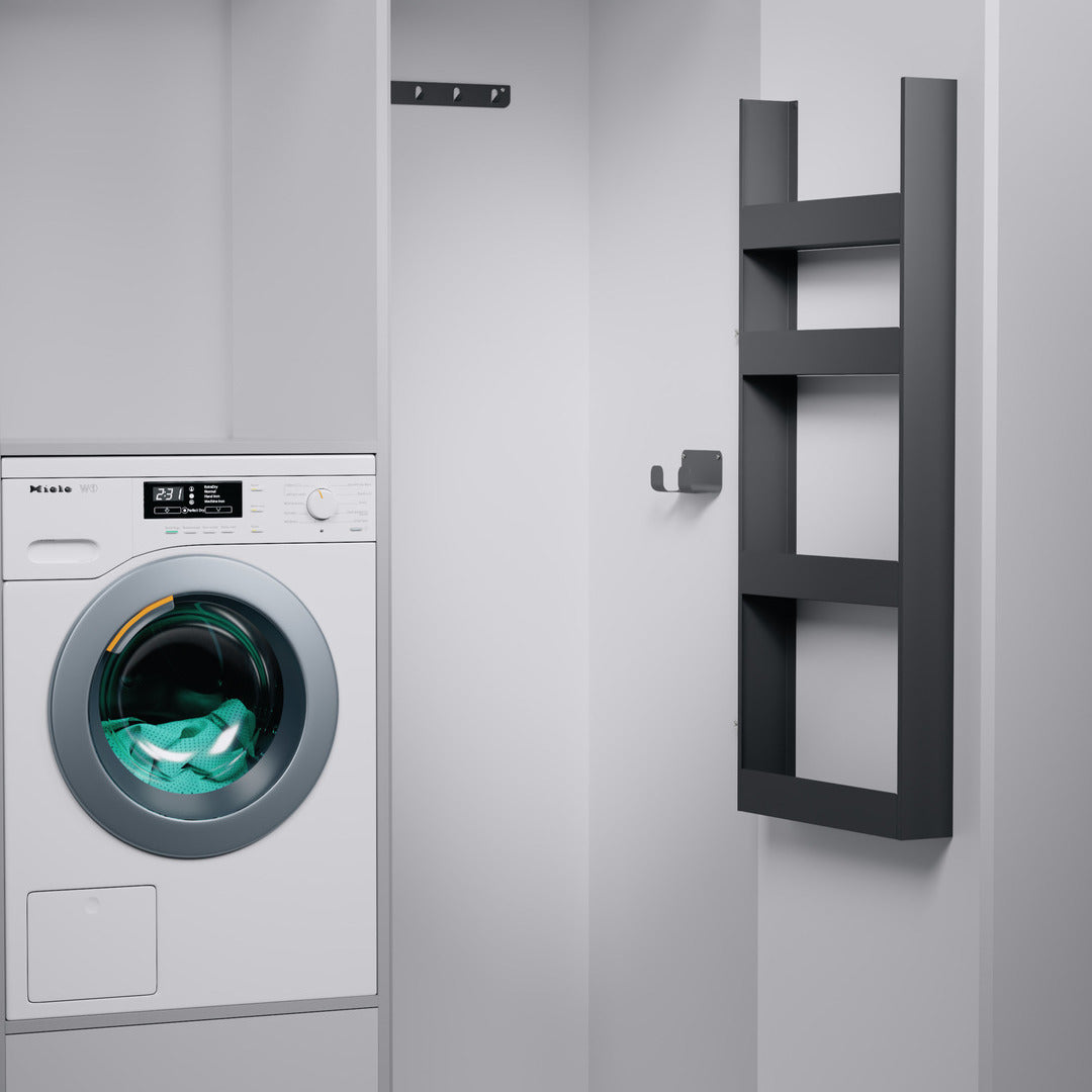 Eine moderne Waschküche mit einer Frontlader-Waschmaschine und Handtüchern im Inneren. An den minimalistischen grauen Wänden befinden sich ein schwarzes Wandregal, ein Wandhaken und das seitliche EasyRack von K-Line für effiziente, intelligente Aufbewahrung.