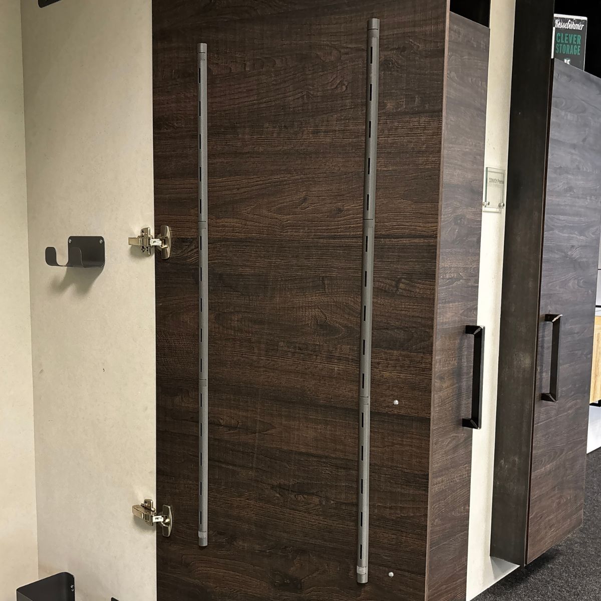 Eine Schranktür aus dunklem Holz mit Metallscharnieren ist geöffnet und zeigt zwei vertikale seitliche Befestigungsleisten von K-Line. An der Wand daneben befinden sich ein Metallhaken und ein weiterer Schrank mit schwarzen Griffen, die auf ein praktisches Ordnungssystem hinweisen.