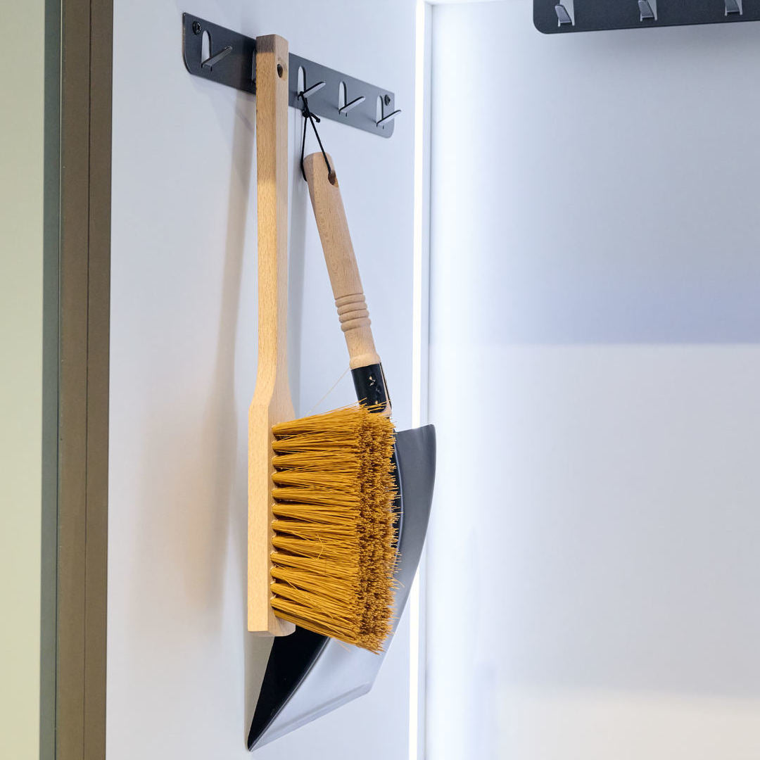 Ein Besen und eine Kehrschaufel mit Holzgriffen hängen platzsparend von der K-Line Seite Hakenleiste - seitlich anschraubbar an einer weißen Wand, neben einer offenen Tür.