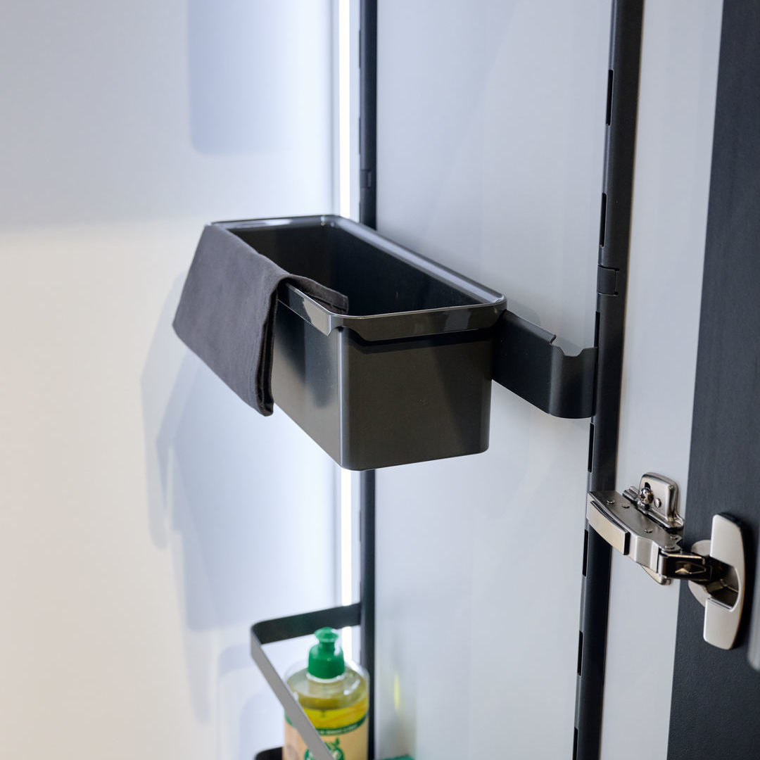 Ein schwarzer rechteckiger Behälter aus der K-Line Trägerleiste mit Box, drapiert mit dunklem Stoff, ist an einer Schranktür für Küchenschrank organisieren montiert. Darunter hält ein Regal eine Flasche mit grünem Deckel am metallischen Scharnier und Riegel.