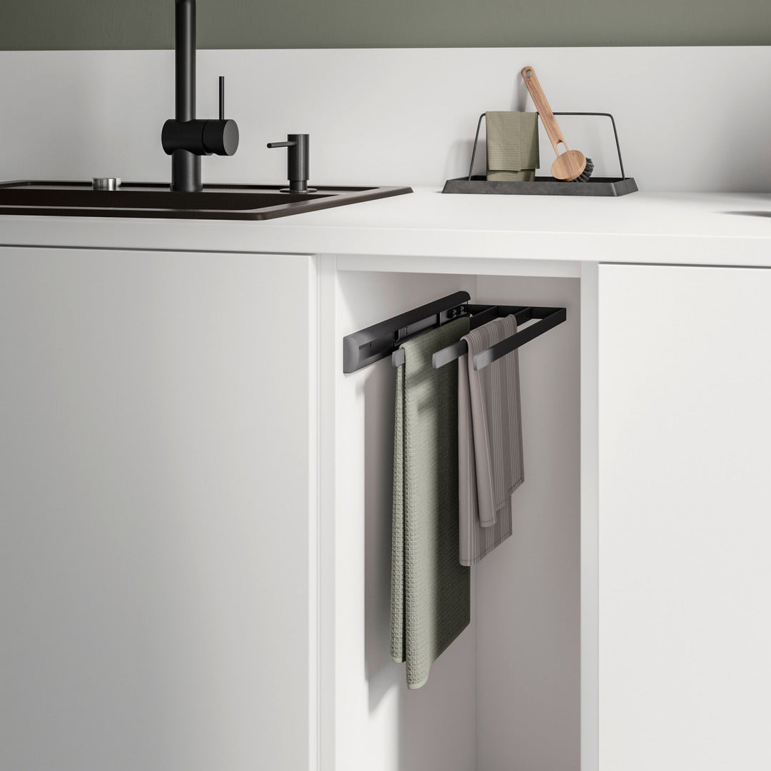 Eine moderne Küche mit einer weißen Schranktür, die einen 3-armigen Handtuchauszug offenbart. Darüber befinden sich ein schwarzes Waschbecken und ein schwarzer Wasserhahn neben einem Geschirrtrockengestell und einer Schrubbbürste auf der Arbeitsplatte.