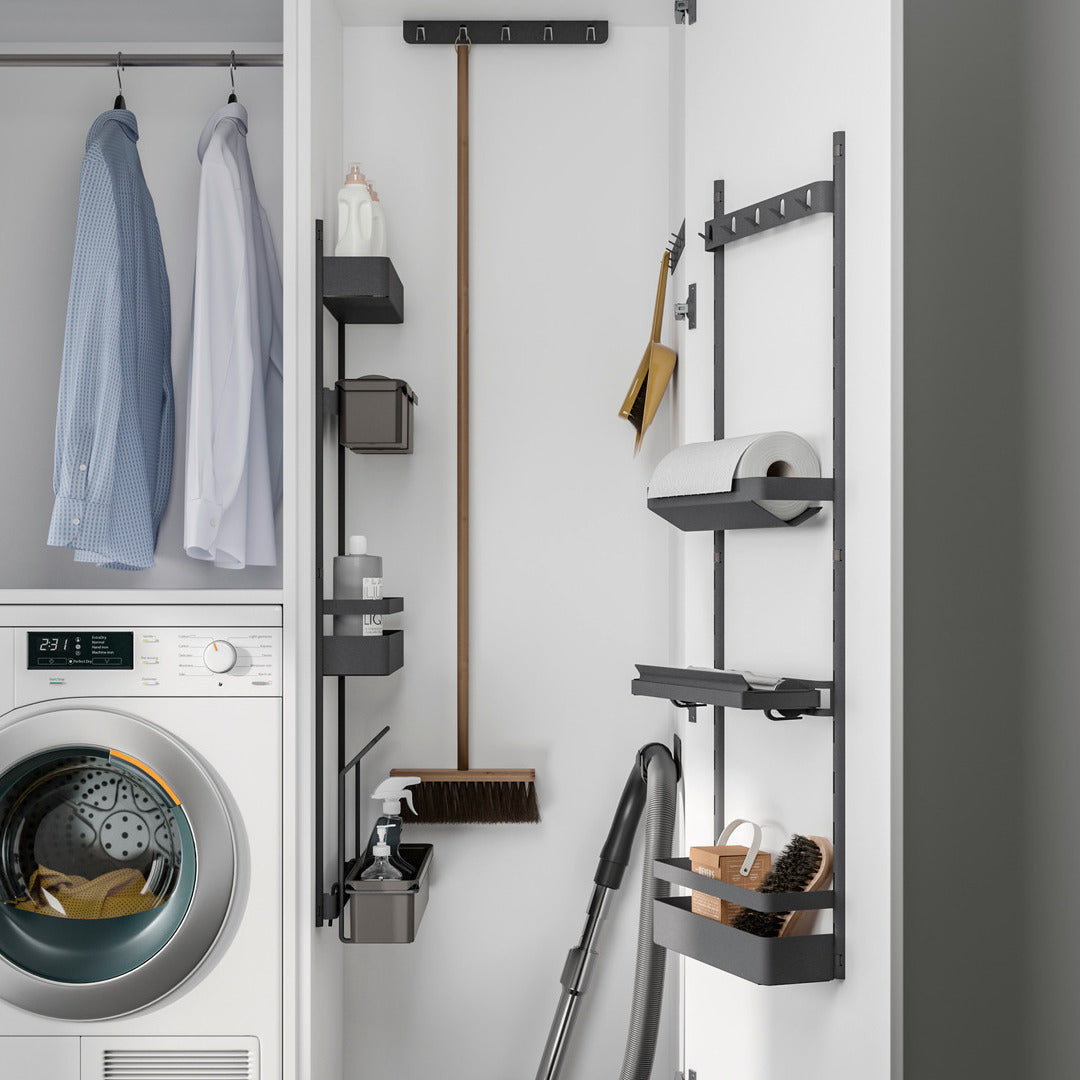Ein kompakter Wäscheschrank verfügt über eine Waschmaschine, hängende Hemden, Regale mit Reinigungsmitteln, einen Staubsauger, einen Besen und eine K-Line-Seitenablage mit Reling zur organisierten Aufbewahrung von Haushaltsgegenständen und Papierhandtüchern.
