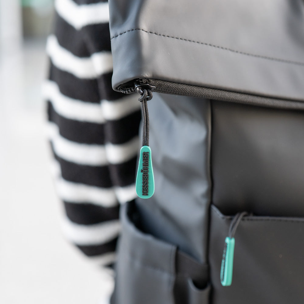 Nahaufnahme des Kesseböhmer Rucksacks, eines schwarzen Rucksacks mit türkisfarbenen Reißverschlüssen und dezentem Aufdruck, getragen über einem schwarz-weiß gestreiften Pullover. Ideal für den Transport von Mahlzeiten oder einer Brotdose an stressigen Tagen.