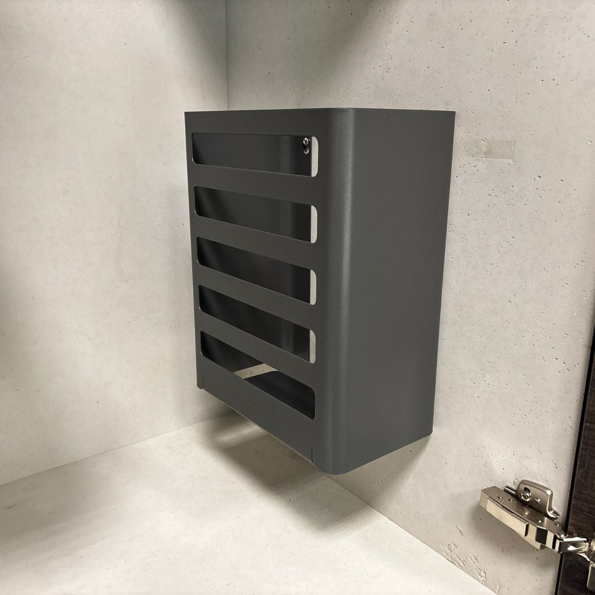 Eine rechteckige, dunkelgraue K-Line Utensilienbox aus Metall mit horizontalen Schlitzen ist an der Innenseite eines hellen Schranks in der Nähe eines silbernen Scharniers angebracht.