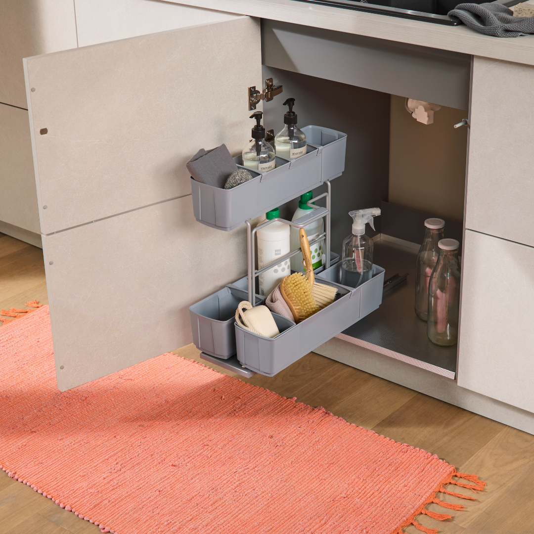 Offener Küchenschrank mit dem cleaningAGENT - Putzschrank Organizer & Putzmittelauszug mit Reinigungsmitteln, Schwämmen und Seifenspendern. Darin stehen Glasflaschen. Ein korallenroter Teppich liegt auf dem Holzboden davor.